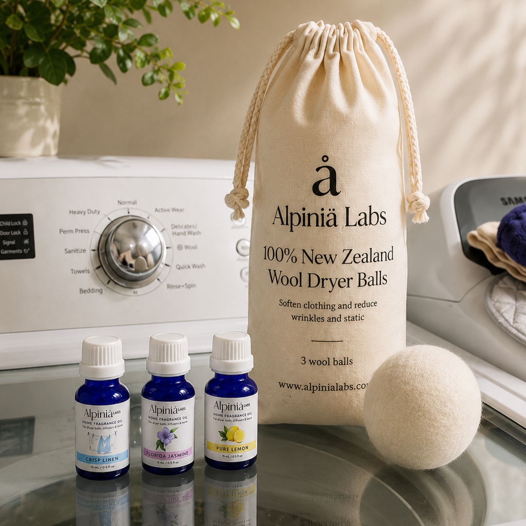 Alpínia Labs Wool Dryer Balls set displayed on a glass shelf in front of a washing machine, featuring a beige drawstring bag, three small blue essential oil bottles, and a white wool dryer ball.